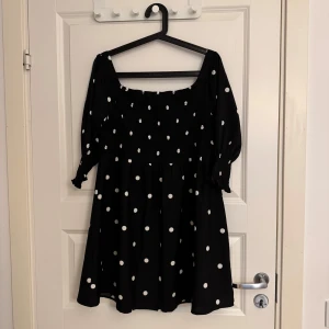 Svart prickig kortklänning Boohoo - Superfin svart kortklänning från Boohoo med vita prickar, smockad överdel och trekvartsärmar med resår och liten volang. Klänningen har en söt, luftig passform och fyrkantig ringning. Perfekt för dig som gillar lekfulla mönster och trendiga detaljer.