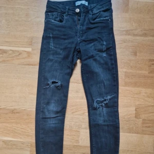 Svarta skinny jeans från Zara - Säljer ett par svarta skinny jeans från Zara med slitna detaljer på benen och fransade benslut. Jeansen har klassisk femficksdesign, hög midja och dragkedja med knapp. Perfekta för en cool och avslappnad look.