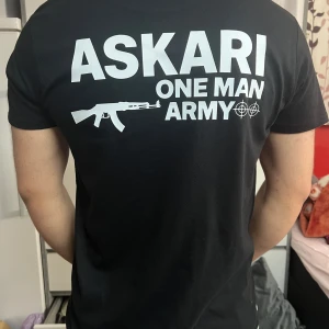 Svart ASKARI t-shirt med tryck - Svart t-shirt från ASKARI med vit text och motiv på både bröstet och ryggen. Stort tryck på ryggen med texten 'ASKARI ONE MAN ARMY' och en gevärsgrafik. Klassisk passform och rund hals. Perfekt för dig som gillar statement-plagg.