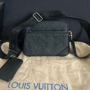 Svart axelväska i konstläder - Snygg svart axelväska liknade Louis Vuitton i präglat konstläder med LV-monogram. Väskan har två stora fack med dragkedja, justerbar axelrem och en liten avtagbar ficka på remmen. Perfekt storlek för mobilen, plånboken och andra essentials. Helt ny