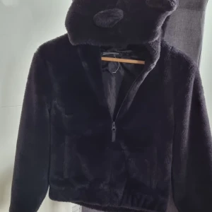 Svart teddyjacka med öron från Destination - Mysig svart teddyjacka från Destination i storlek 152. Jackan har huva med söta öron, dragkedja framtill och är helfodrad. Perfekt för dig som vill ha en varm och cool look med lite extra attityd.