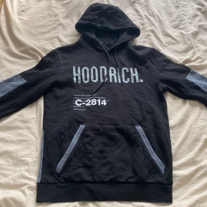 Svart/grå Hoodrich huvtröja - Säljer en svart grå hoodie från HOODRICH med grå detaljer på ärmar och nederkant. Stor logga framtill och världskarta med tryck på ryggen. Huva med snörning och stor magficka. Perfekt streetwear-vibe för dig som gillar snygga prints.