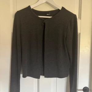 Mörkgrå kofta från Gina Tricot - En stilren mörkgrå kofta från Gina Tricot med rund halsringning och en enkel knapp framtill. Koftan är långärmad och har en klassisk, rak passform. Perfekt att slänga över en topp för en chill look. Jag har oftast storlek xs/s men i detta fallet var den väldigt liten i passform, alltså gick jag upp en storlek. Den har använd ett fåtal gånger och har inga defekter. Den säljs inte på hemsidan längre alltså vet jag inte vad ny priset är.