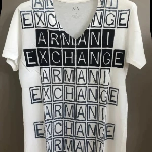 Vit t-shirt Armani Exchange tryck - Vit t-shirt från Armani Exchange med svart grafiskt tryck över hela framsidan där det står 'Armani Exchange' i rutmönster. T-shirten har v-ringning och korta ärmar, perfekt för en clean och modern streetstil.