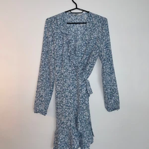 Blå blommig omlottklänning från ONLY - Supersöt blå kort omlottklänning från ONLY med vitt blommönster, långa ärmar och volangkant nertill. Knyt i midjan för en snygg passform. Perfekt för exempelvis skolavslutning, midsommar eller en vanlig sommardag 