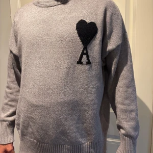 Grå stickad tröja från Ami Paris - Säljer en grå stickad tröja från Ami Paris med svart hjärta och A-logga på bröstet. Tröjan har rund hals, ribbade muddar och en loose passform. Perfekt för dig som gillar stilren design med en twist.