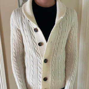 Vit kabelstickad kofta från Buddha - Snygg vit kabelstickad kofta från Buddha med stora knappar och bred krage. Koftan har lång ärm och ribbade muddar, perfekt för lager på lager. Klassisk och stilren look som funkar till jeans eller kjol.