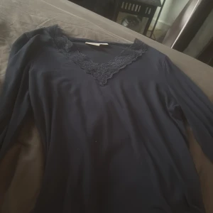 Mörkblå långärmad topp med spets - Snygg mörkblå långärmad topp med v-ringning och dekorativ spets runt halsen. Perfekt för dig som gillar en stilren look med lite extra detalj. Mjuk och skön, passar till både jeans och kjol.
