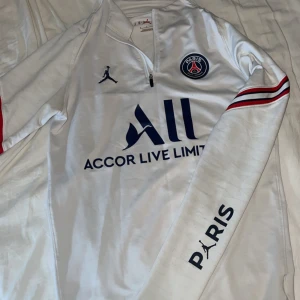 Vit PSG långärmad fotbollströja Jordan - Snygg vit långärmad PSG-tröja från Jordan med blå och röda detaljer på ärmarna. Halv dragkedja framtill, Paris Saint-Germain-logga på bröstet och tryck i marinblått. Materialet är lätt och sportigt, perfekt för träning eller match.storlek s men är rätt så stor i storleken.