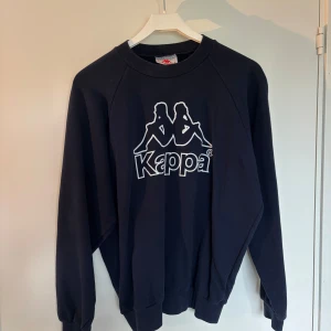 Svart sweatshirt från Kappa - Säljer en svart sweatshirt från Kappa med stort vitt logotryck på bröstet. Tröjan har rund hals, långa ärmar och ribbade muddar vid ärmslut och nederkant. Perfekt för dig som gillar sportig och avslappnad stil.