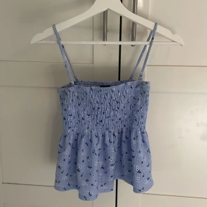 Blommig blå linnetopp från Gina Tricot - Supersöt ljusblå linnetopp från Gina Tricot med smala axelband och blommigt mönster i vitt och mörkblått. Toppen har smockad överdel och volangig nederkant, perfekt för en somrig vibe. Materialet är lätt och luftigt, vilket gör den extra skön under varma dagar.