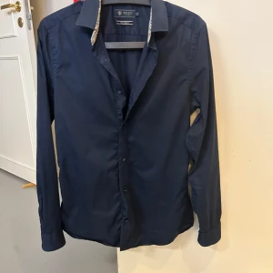 Mörkblå skjorta Riley Slim Fit - Snygg mörkblå skjorta från Riley i slim fit-modell. Skjortan har klassisk krage, knäppning framtill och långa ärmar. Perfekt för dig som gillar en stilren och tajt passform.