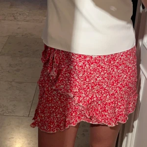 Röd blommig volangkjol med knyt - Superfin röd kortkjol med vita blommor och volangkant. Kjolen har omlottmodell och knyt i sidan, vilket ger en söt och somrig vibe. Perfekt för varma dagar och lätt att matcha med en enkel topp.