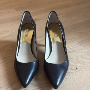 Svarta pumps från Michael Kors - Klassiska svarta pumps från Michael Kors i skinn med spetsig tå och elegant låg klack. Snygg gulddetalj baktill och lyxig guldfärgad innersula. Perfekta för dig som gillar stilrena och tidlösa skor.