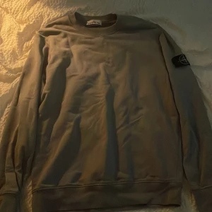 Grå sweatshirt från Stone Island - Säljer en clean grå sweatshirt från Stone Island med klassisk patch på ärmen. Tröjan har rund hals, långa ärmar och är gjord i mjuk bomull. Perfekt för dig som gillar stilrena och ikoniska plagg med streetkänsla. Säljer pga att den inte passar längre.  tröjan är äkta ni kan kolla certilogo koden inga skambud! 