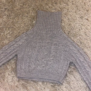 Grå stickad polotröja Gina Tricot - Supermysig grå stickad polotröja från Gina Tricot i croppad modell. Tröjan har flätstickat mönster, hög polokrage och långa ärmar. Perfekt för kalla dagar och enkel att styla med jeans eller kjol.