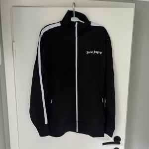 Palm Angels svart träningsset - Säljer ett svart träningsset från Palm Angels med vita ränder längs ärmar och ben. Jackan har dragkedja framtill och logga på bröstet, byxorna har samma logga och dragkedjor vid bensluten. Snygg och sportig look med klassiska detaljer.