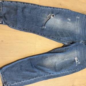 Blå baggy jeans med slitningar - Säljer ett par blå jeans med coola slitningar och ljusa tvättade partier. Byxorna har en baggy passform och klassisk femficksdesign. Perfekta för en avslappnad streetstil och tillverkade i mjukt denimtyg.