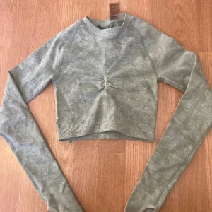 Grön croppad tröja från Gymshark - Snygg ljusgrön croppad tröja från Gymshark med lång ärm och coola cut-outs i ryggen. Tröjan har ett diskret camo-mönster, ribbade muddar och en liten rynkdetalj framtill. Perfekt för träning eller chill dagar.