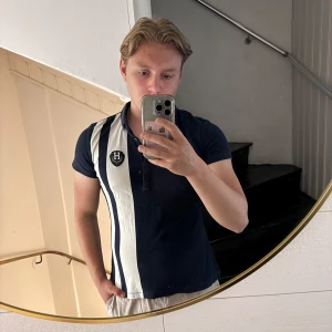 Tommy Hilfiger pikétröja marinblå/vit - Snygg pikétröja från Tommy Hilfiger i marinblått och vitt med breda vertikala ränder och broderad logga på bröstet. Klassisk krage, kortärmad och knäppning framtill. Perfekt för en sportig och stilren look. Sitter som S