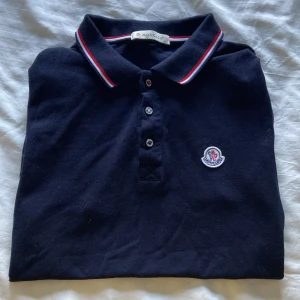 Mörkblå Moncler pikétröja S - Snygg mörkblå pikétröja från Moncler. Klassisk krage med röd, vit och blå rand, tre knappar framtill och broderad Moncler-logga på bröstet. Perfekt för dig som gillar stilrena vibes.