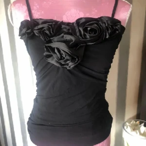 Svart linnetopp med stora rosor - Supersnygg svart linnetopp med smala axelband och stora tygrosor framtill. Toppen är figurnära och har en cool, lite glansig känsla upptill tack vare rosdetaljerna. Perfekt för dig som vill sticka ut med en unik och trendig look.
