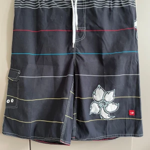 Svarta randiga badshorts med blomma - Svarta badshorts med tunna ränder i vitt, rött, blått och gult samt stora vita blommor som detalj. Shortsen har snörning i midjan och en praktisk ficka med lock på sidan. Perfekta för stranden eller poolen, med en avslappnad och cool vibe.