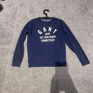 Mörkblå sweatshirt från GANT - Snygg mörkblå sweatshirt från GANT med vit tryckt text på bröstet. Tröjan har rund hals, långa ärmar och ribbade muddar. Perfekt för en chill och sportig look. Tillverkad i mjuk bomull för extra komfort.