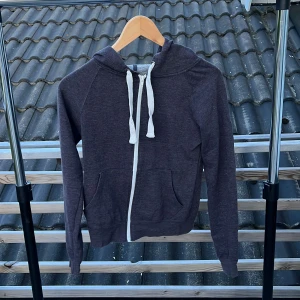 Mörkgrå hoodie från Atmosphere - Mysig mörkgrå hoodie från Atmosphere med vit dragkedja och snören i huvan. Tröjan har två fickor framtill och är långärmad. Perfekt för chill dagar eller när du vill ha en enkel och stilren look.