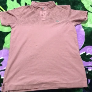 Rosa mönstrad t-shirt från Lacoste - Säljer en rosa t-shirt från Lacoste med korta ärmar och diskret rutmönster upptill. Tröjan har ståkrage med knappar och den klassiska krokodilloggan broderad på bröstet. Tillverkad i mjuk bomull, perfekt för en chill och stilren look.