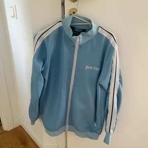 Palm Angels blå  - Snygg ljusblå vindjacka från Palm Angels med vita och svarta stripes längs ärmarna. Jackan har dragkedja framtill, ståkrage och logga på bröstet. Perfekt för dig som gillar streetwear och vill sticka ut med en fräsch färg.
