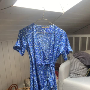 Blå omlottklänning med mönster - Kort blå omlottklänning med grön/vita detaljer. Använd en gång. 