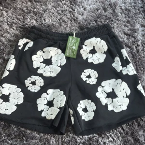 Svarta shorts från Denim Tears XL - Svarta shorts från Denim Tears i storlek XL med unikt blommigt mönster i vitt. De har elastisk midja med snörning och är tillverkade i mjukt bomullsmaterial. Perfekta för en chill och trendig sommarstil.
