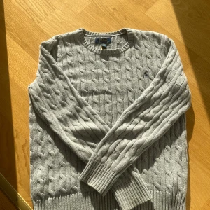 Grå kabelstickad tröja Polo Ralph Lauren - Kabelstickad grå tröja från Polo Ralph Lauren med klassisk rund halsringning och diskret broderad logga på bröstet. Tröjan har långa ärmar och ribbade muddar för en snygg och tidlös look. Perfekt för lager-på-lager-stil.