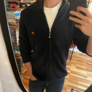 Mörkblå helzip  - Mörkblå ribbstickad kofta från Denim Culture med hel dragkedja och ståkrage. Liten orange broderad logga på bröstet ger en clean look. Perfekt till jeans eller chinos för en chill och stilren vibe. Köpt på plick för ungefär ett halvårsen, aldrig använd. 