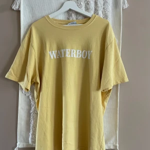 Gul Nikben t-shirt med tryck - Gul t-shirt från Nikben i 100% ekologisk bomull. Framsidan har vit text 'WATERBOY' och baksidan ett stort konstnärligt tryck med en sjöjungfru och pelare. Klassisk passform och rund hals, perfekt för dig som gillar unika prints och chill vibe.