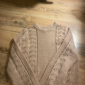 Beige stickad tröja med hålmönster - Säljer en beige stickad tröja med snyggt hålmönster längs ärmar och framstycke. Tröjan har rund halsringning och långärmade ärmar. Perfekt för dig som gillar en mysig och trendig look under kyliga dagar.