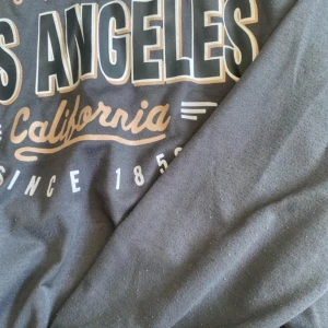 Svart Los Angeles sweatshirt - Cool svart sweatshirt med tryck 'Los Angeles California West Coast' i beige och vitt framtill. Tröjan har rund halsringning och långa ärmar. Perfekt för dig som gillar streetstyle och vill ha en avslappnad look.