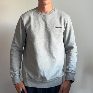 Grå sweatshirt från Carhartt - Snygg grå sweatshirt från Carhartt med minimalistisk logga på bröstet. Tröjan har rund halsringning, långa ärmar och ribbade muddar. Perfekt för en clean och avslappnad stil.