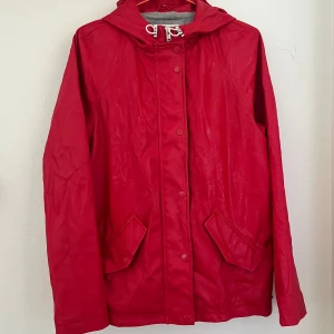 Röd regnjacka med huva och snörning - Snygg röd regnjacka med huva och fodrad insida. Jackan har två stora fickor framtill med lock och stängs med knappar. Perfekt för regniga dagar och enkel att styla till jeans eller sneakers. 