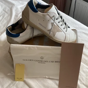 Vita Golden Goose sneakers med blå detalj - Golden Goose sneakers i vitt skinn med grå snörning, beige mockastjärna på sidan och blå detalj vid hälen. Klassisk rund tå och platt sula. Snygg streetstil med ikonisk GGDB-stjärna och vintagekänsla. Kommer med originalkartong och dustbag.