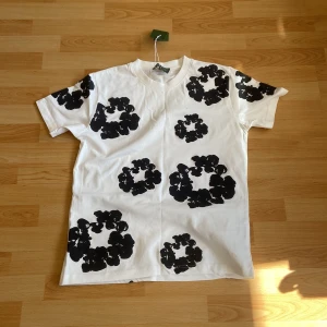 Vit t-shirt med svarta blommor Denim Tears - Vit t-shirt från Denim Tears med stora svarta blommiga tryck över hela plagget. Klassisk passform med rund hals och korta ärmar. Tillverkad i mjuk bomull, perfekt för dig som gillar streetwear med unik design.
