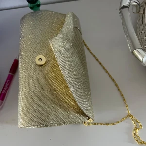Glittrig guldig kuvertväska med kedja - Säljer en snygg kuvertväska i guld med glittrig yta och guldfärgad kedja. Väskan har magnetlås och stilren design, perfekt för att lyfta din outfit. Insidan är svart och väskan är lätt att bära över axeln eller i handen.