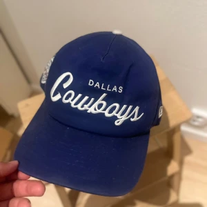 Dallas Cowboys keps Super Bowl XXVII - Blå Dallas Cowboys keps från New Era med broderad Super Bowl XXVII-logga och vita detaljer. Justerbar snapback baktill. Broderad text framtill och coolt broderi på sidan med rosor och romerska siffror. Perfekt för NFL-fans som vill sticka ut.