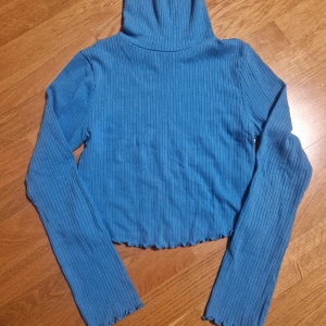 Blå ribbad polotröja från Faster - Säljer en blå ribbad polotröja från Faster i storlek S. Tröjan har lång ärm, hög polokrage och croppad passform med vågig kant nedtill. Perfekt för dig som gillar en enkel men snygg stil med lite edge.