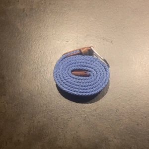 Blått flätat bälte med läderdetaljer - Snyggt blått bälte med flätad design och bruna läderdetaljer vid spännet. Bältet har ett klassiskt metallspänne och känns både stilrent och modernt. Perfekt för att ge outfiten en färgklick och funkar till jeans eller chinos.