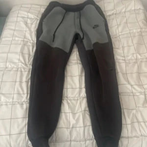 Nike mjukisbyxor i grå och svart S - Nike mjukisbyxor i tvåfärgad design med grå framsida och svarta ben. Byxorna har dragsko i midjan, ribbade muddar och klassisk Nike-logga på låret. Perfekta för chill eller träning, tillverkade i mjukt material för maximal komfort.