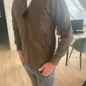 Grön hoodiejacka från C.P. Company - Säljer en grön hoodiejacka stl S från C.P. Company med deras ikoniska goggles i huvan. Jackan har dragkedja framtill, ribbade muddar och två fickor med dragkedja. Perfekt för dig som vill sticka ut med streetwear-vibe och snygga detaljer.