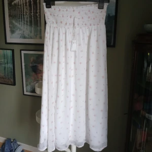 Vit långkjol med små blommor - Superfin vit långkjol med små rosa blommor och smockad midja. Kjolen har knytband med tofsar framtill och ett luftigt, flowy tyg som känns perfekt för varma dagar. Söt och romantisk stil som passar dig som gillar en bohemisk vibe. Köpt för 359 kr.
