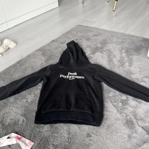 Svart hoodie från Peak Performance - Svart hoodie från Peak Performance med vit logga på bröstet. Klassisk modell med huva och magficka, tillverkad i mjuk bomullsblandning. Perfekt för chill dagar eller när du vill ha en enkel och snygg look.